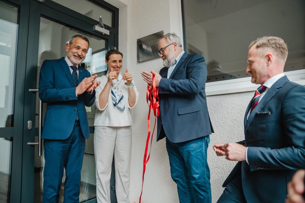 Kobieta i trzech mężczyzn w garniturach stojących razem, uśmiechniętych przed drzwiami do budynku. Jeden z mężczyzn trzyma czerwoną wstęgę.